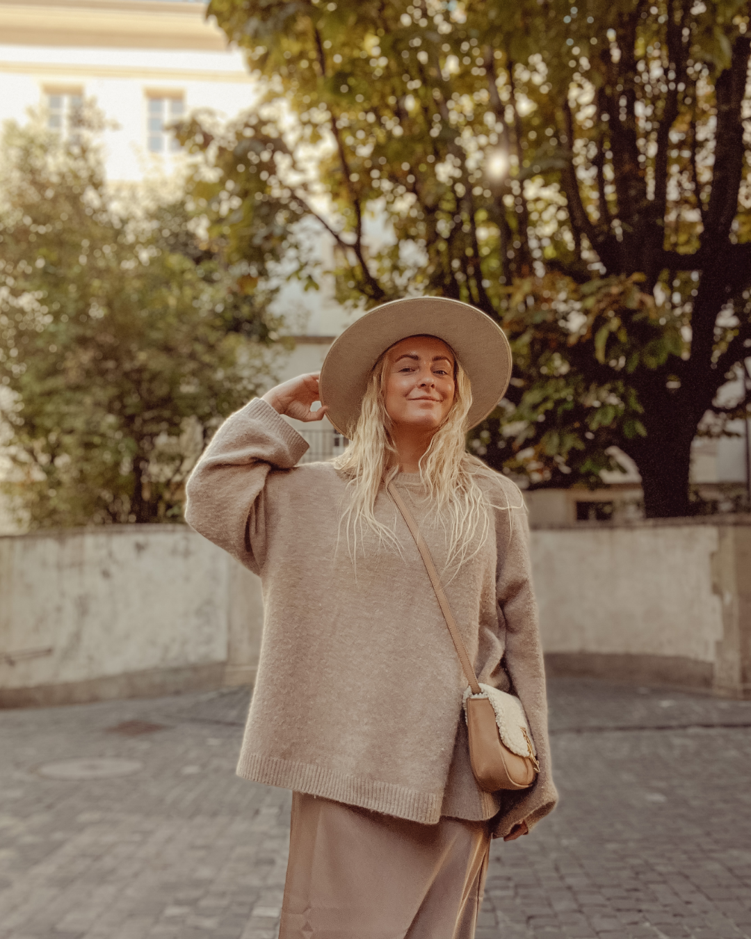 Vaaleahiuksinen nainen beige hattu päässään hymyilee pidellen hatun reunasta kiinni