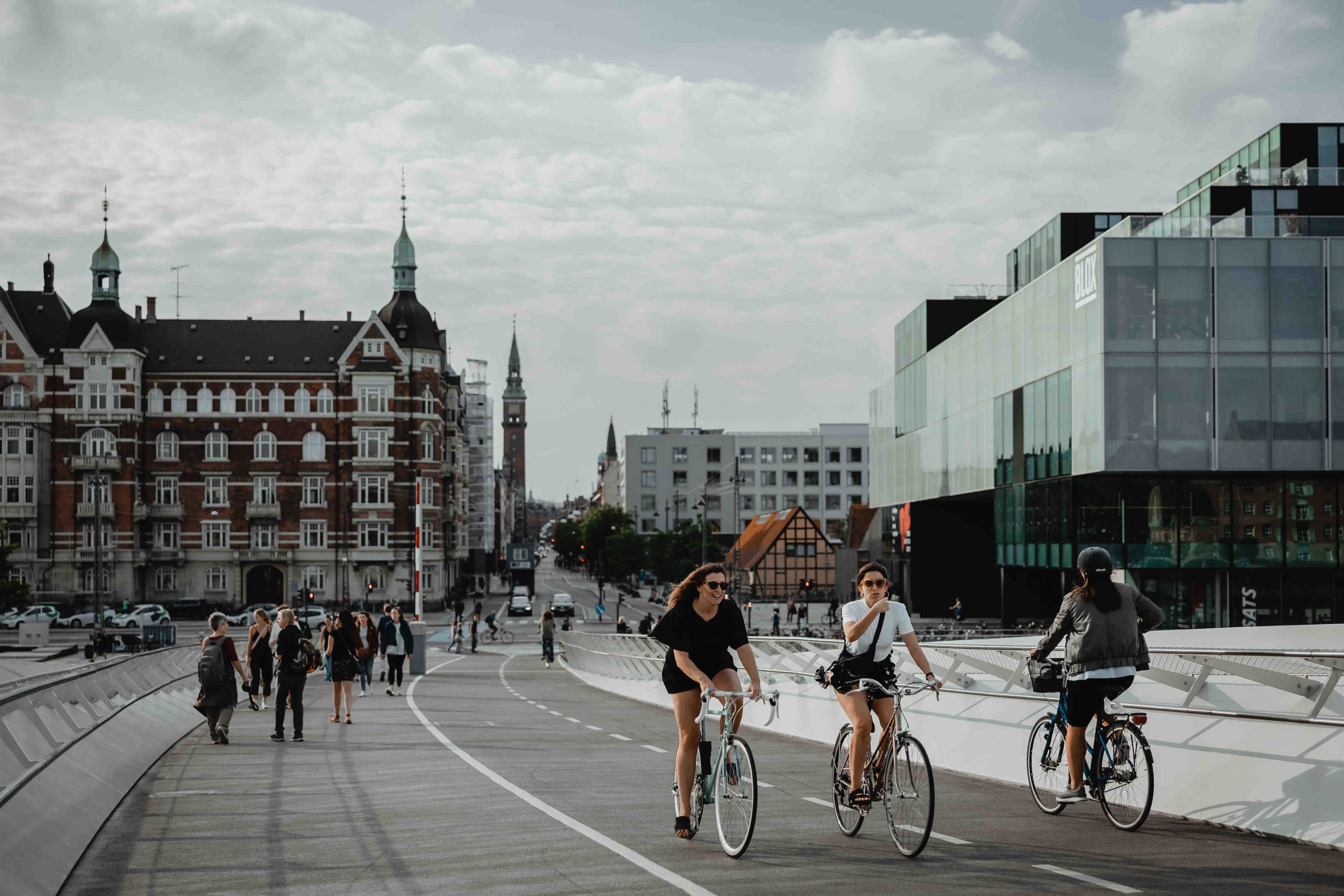 Two girls cycling across a bridge with modernist and traditional buildings in the background.