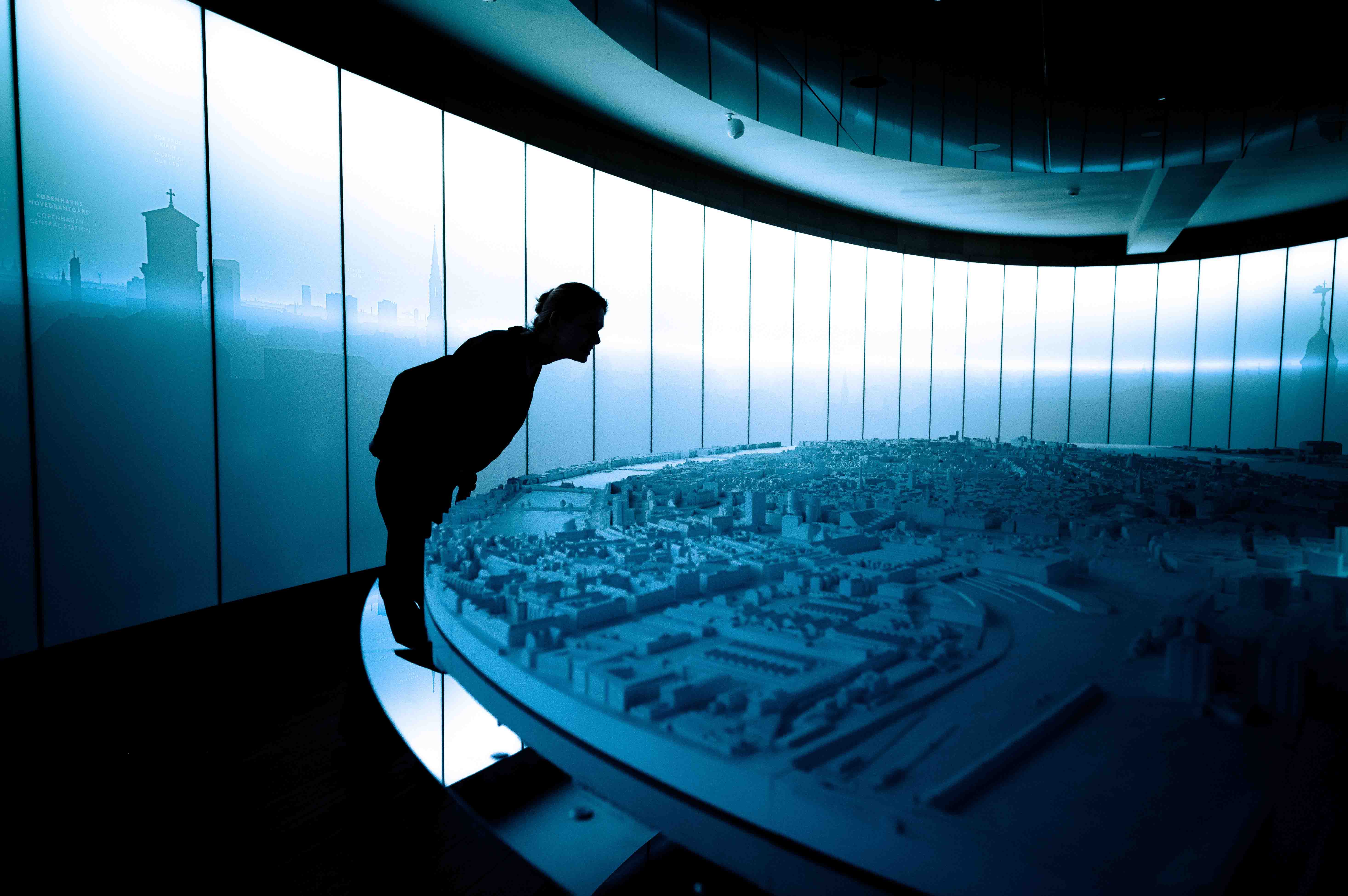A man leaning over a scale model of Copenhagen. Notice the blue light and the silhouettes.