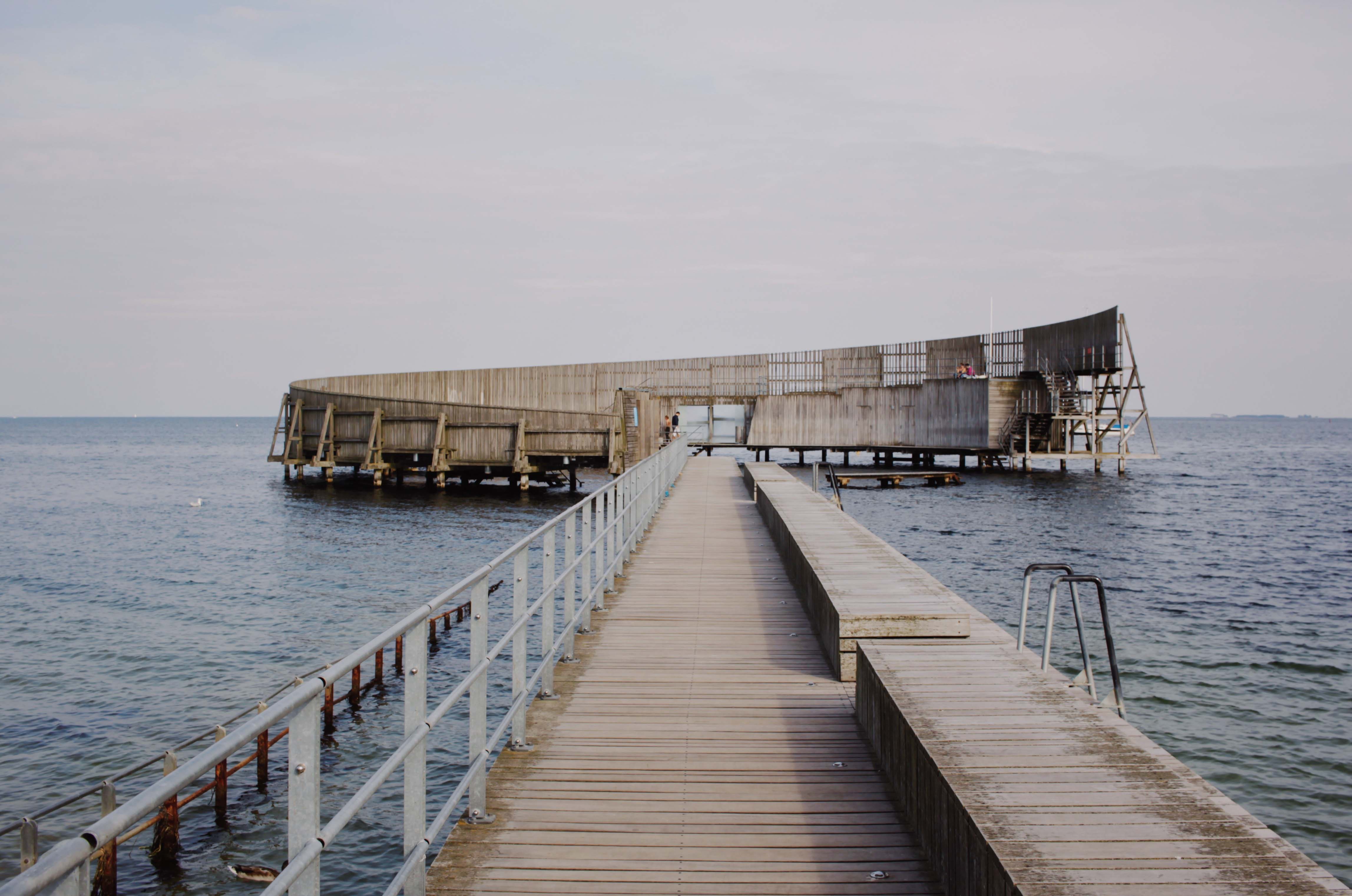 A pier projecting out into the sea and leading to a round, contemporary-looking, wooden bathing house.