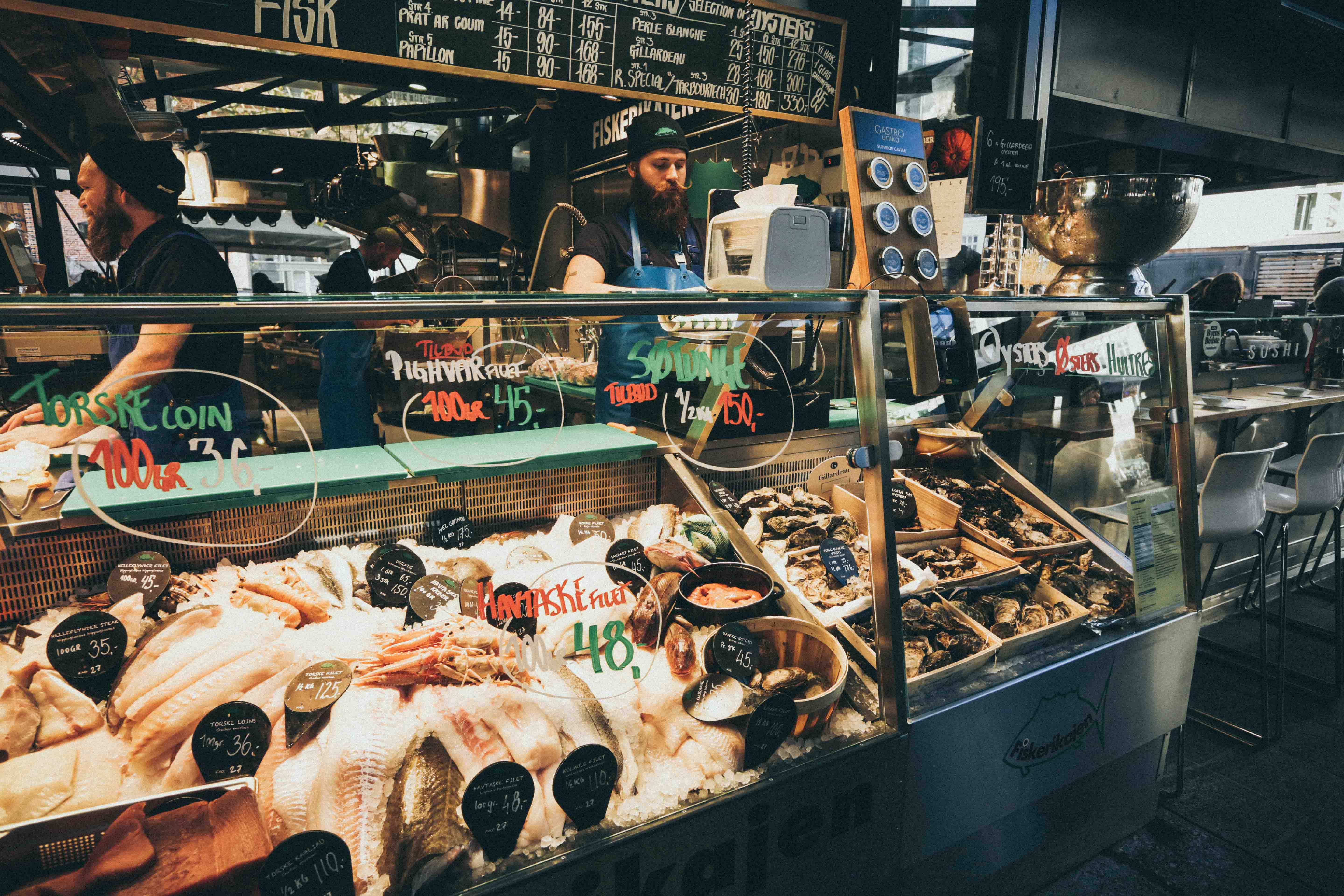 A food market with refrigerated counters filled with fresh fish, with prices written on the glass in red and green. Two bearded men wearing caps standing behind a counter.