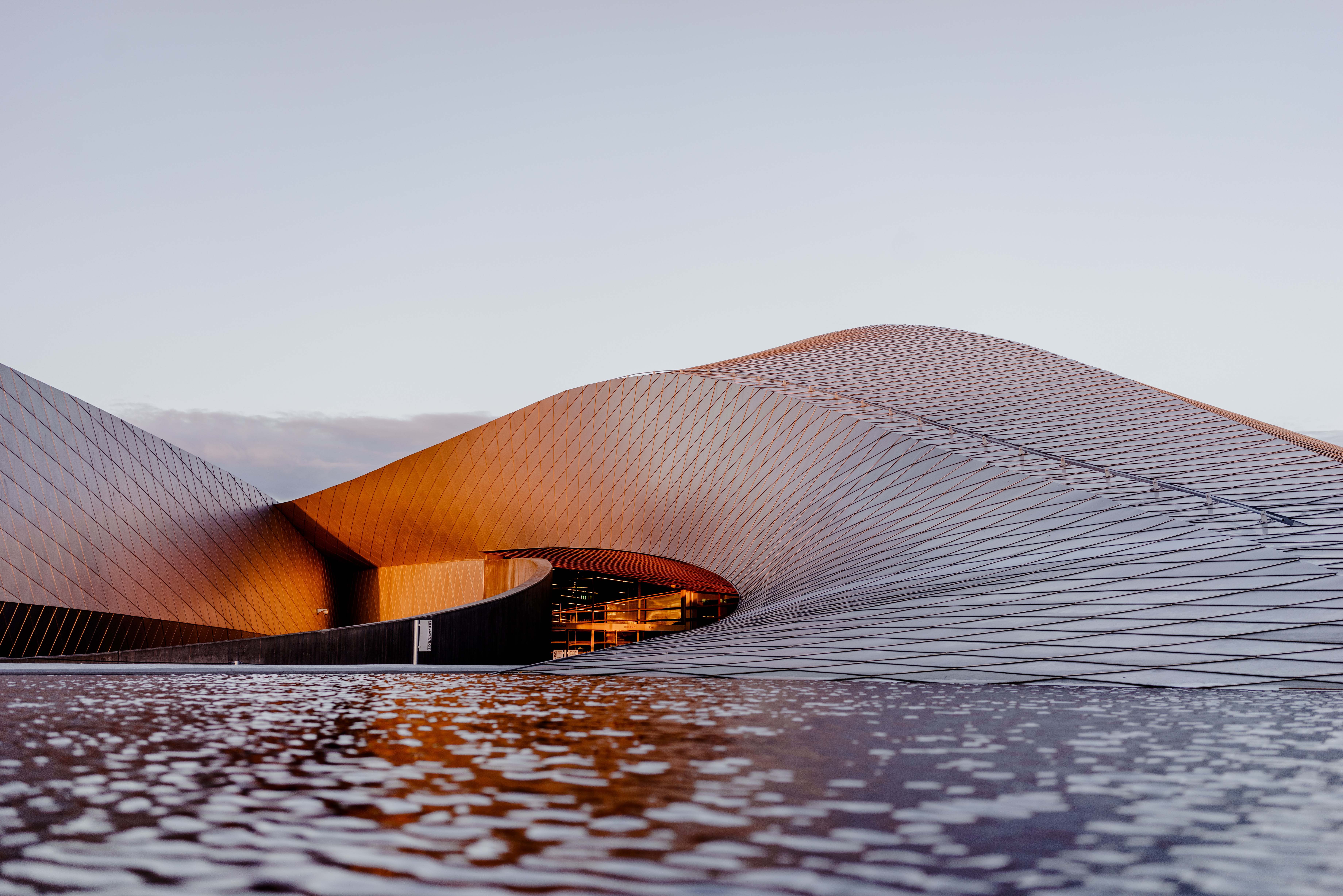 In the foreground, water flows into a modernist building that looks like a sand dune. The sky is blue, but the light gives the impression of sunset.