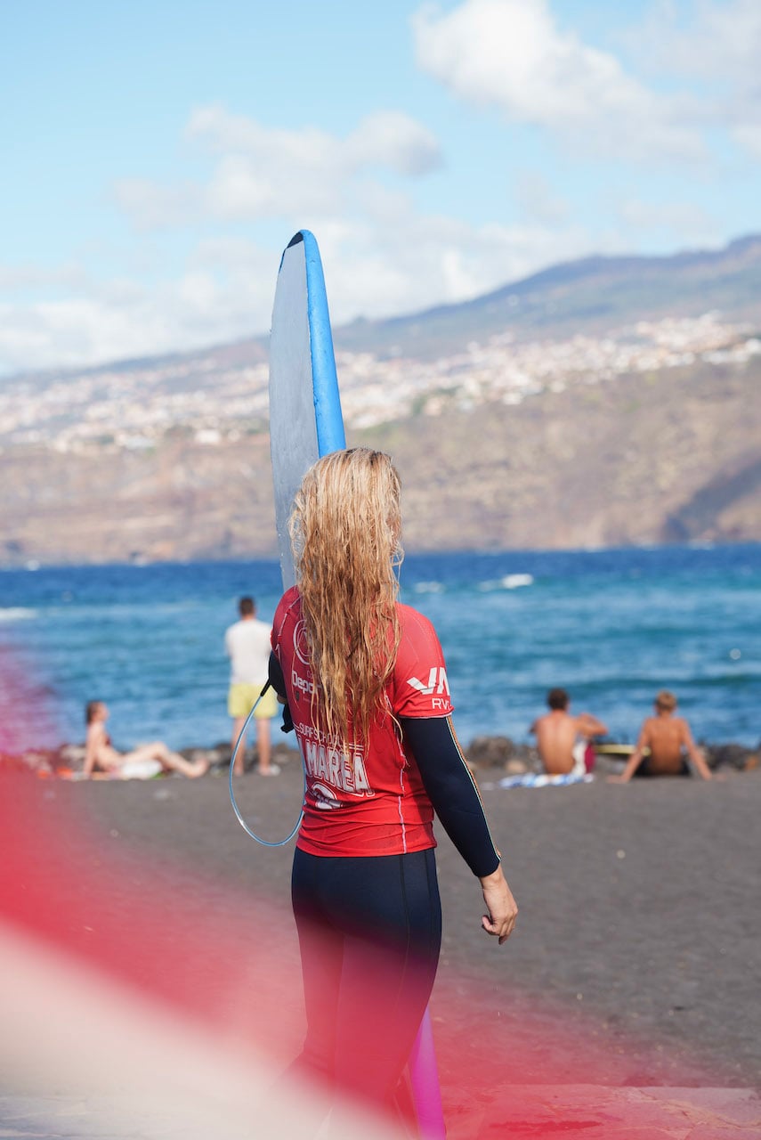 En kvinne med surfeantrekk står på en strand på Tenerife med et surfebrett. 