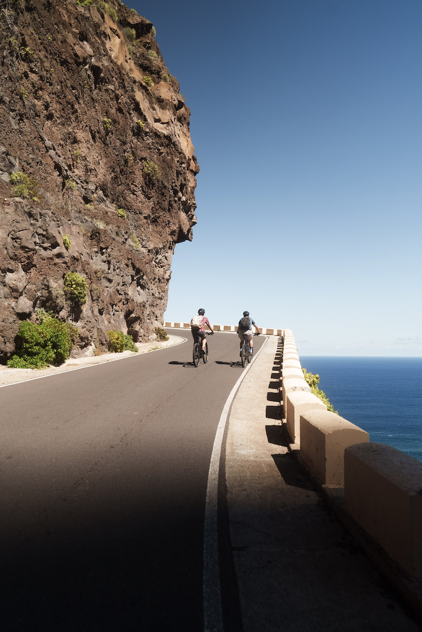 Bikers in the distance going upghill. Mountain wall on one side and the ocean below on the other.