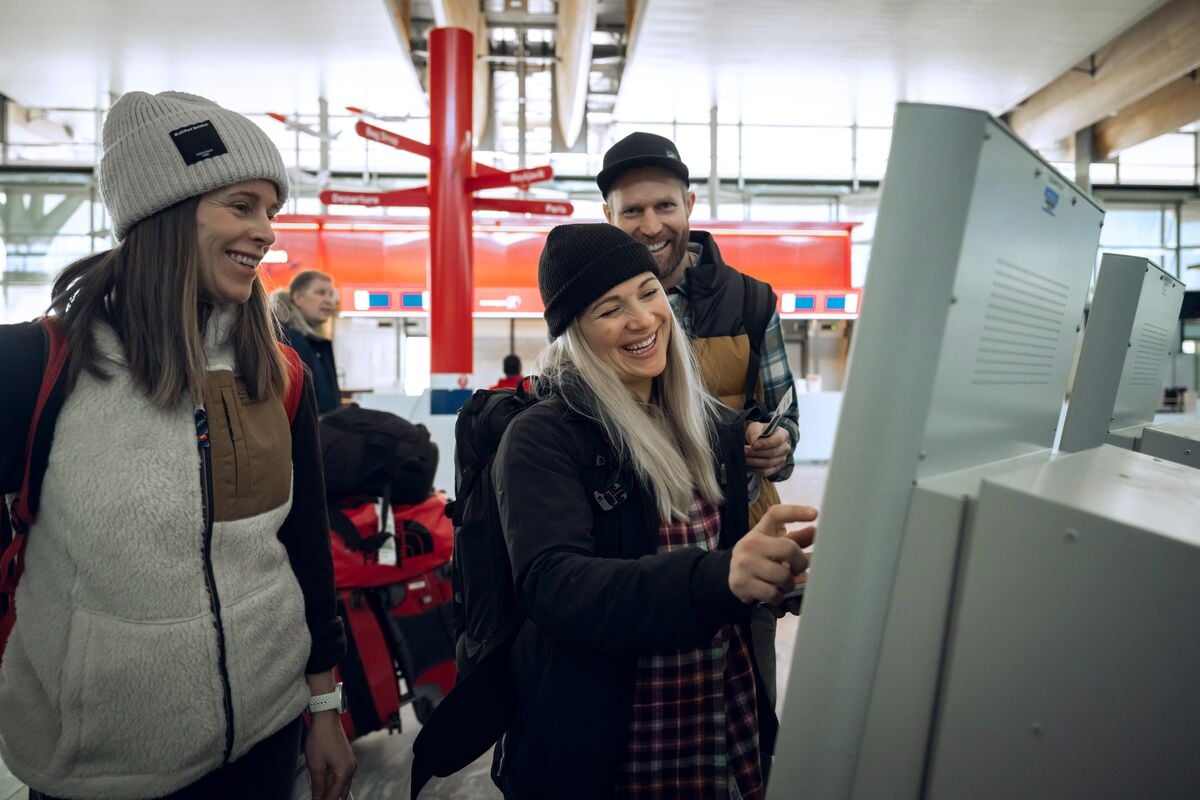 tre venner ler og smiler mens de sjekker inn på automat på flyplassen