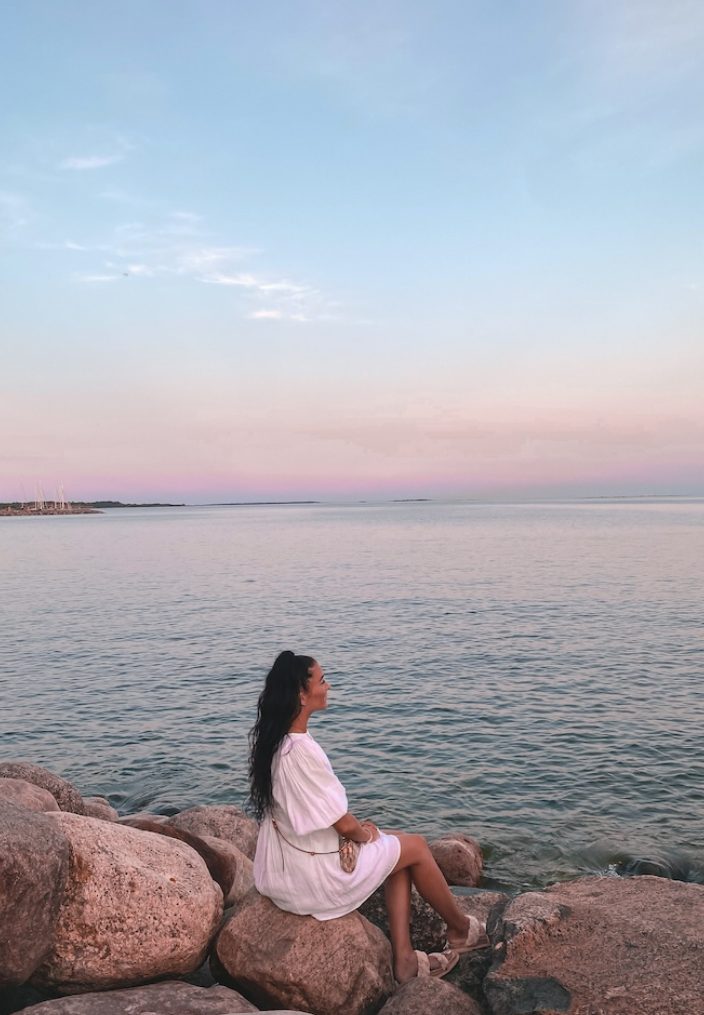 Dark haired woman sitting on large rocks by the ocean with pastel pink sky 