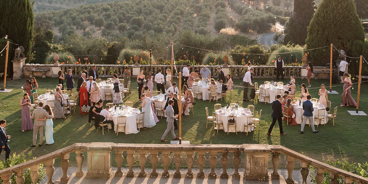En bryllupsfeiring i Toscana med grønne frodige omgivelser, festkledde mennesker og runde bord.