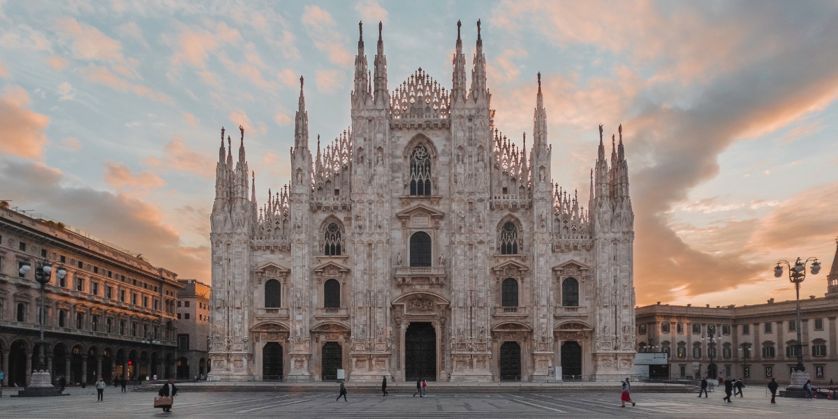 Katedralen i Milano med sina spiror och karaktäristiska form i vit marmor. Platsen framför är tom. Solen är på väg att gå ner bakom kyrkan.