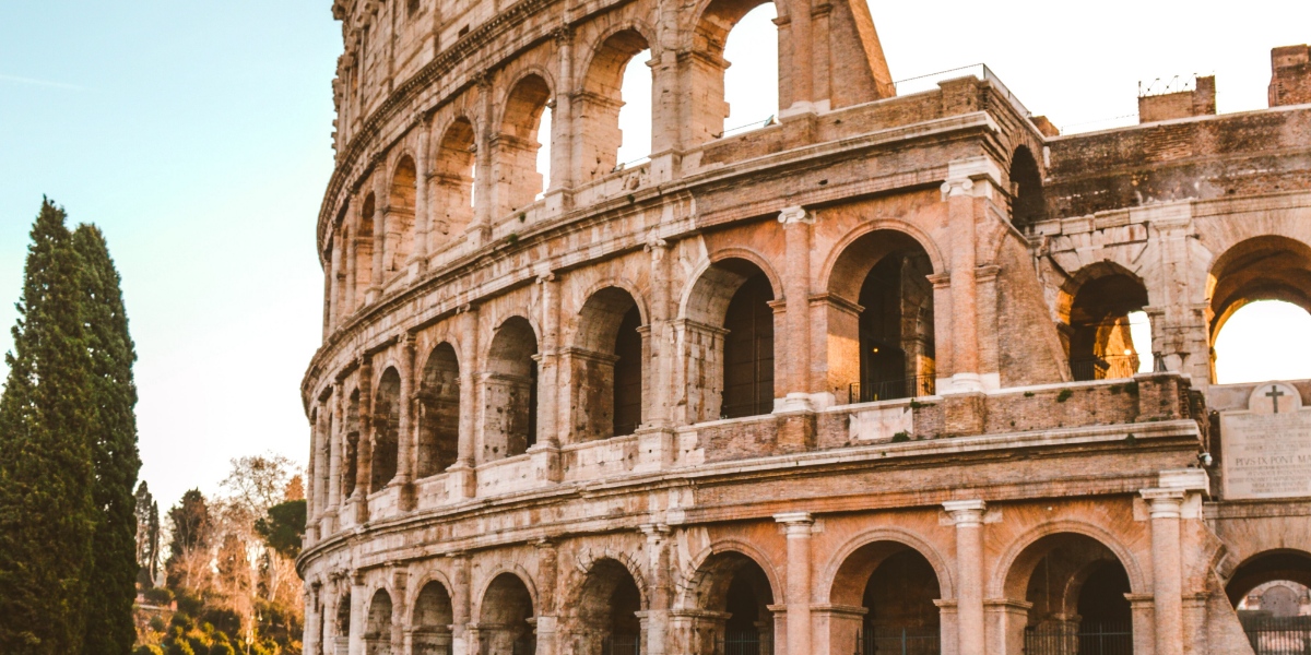 Colosseum med sina många valv i varmt solljus. I bakgrunden ser man en stor tuja och himlen är blå.