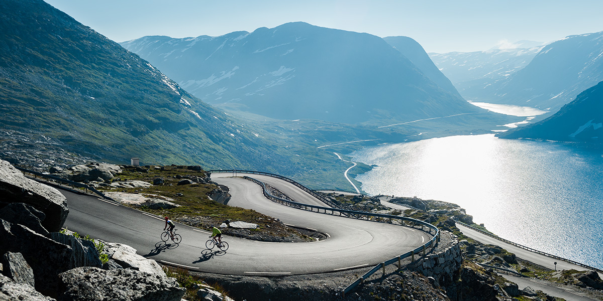 Two bikeriders going uphill on a winding road surrounded by mountains and fjords.