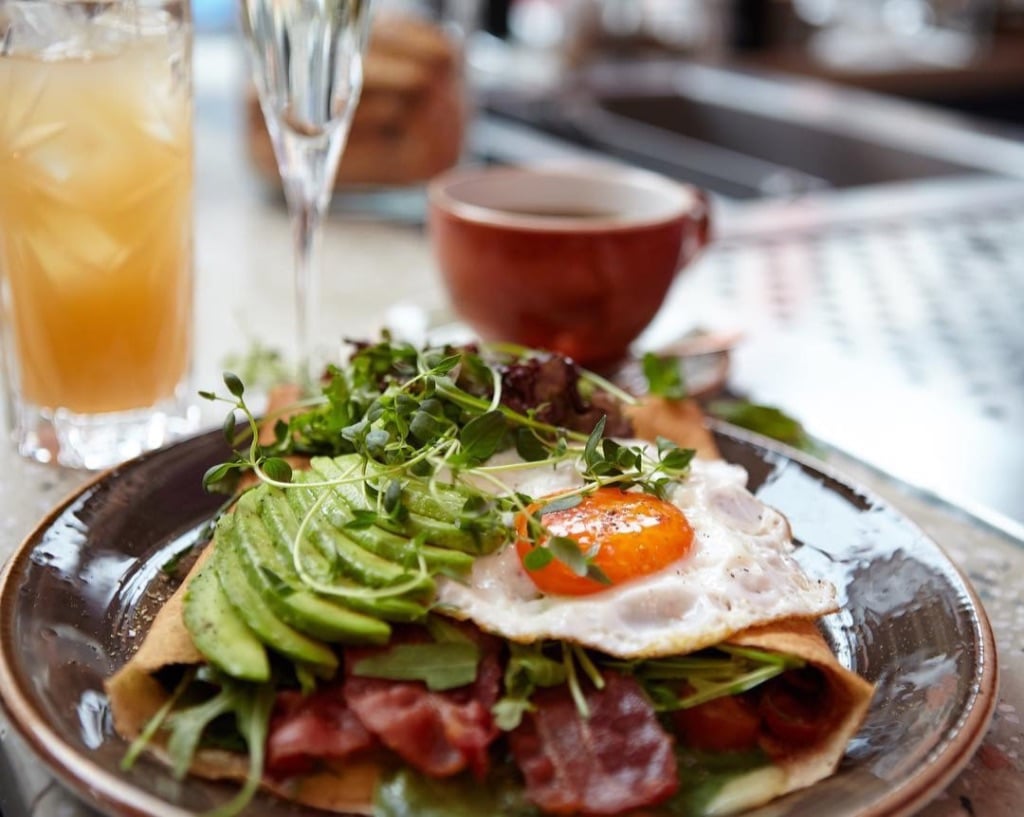 A picture of a crepe with egg, watercress, and avocado with bacon inside. In the background, a coffee cup and orange juice can be seen.