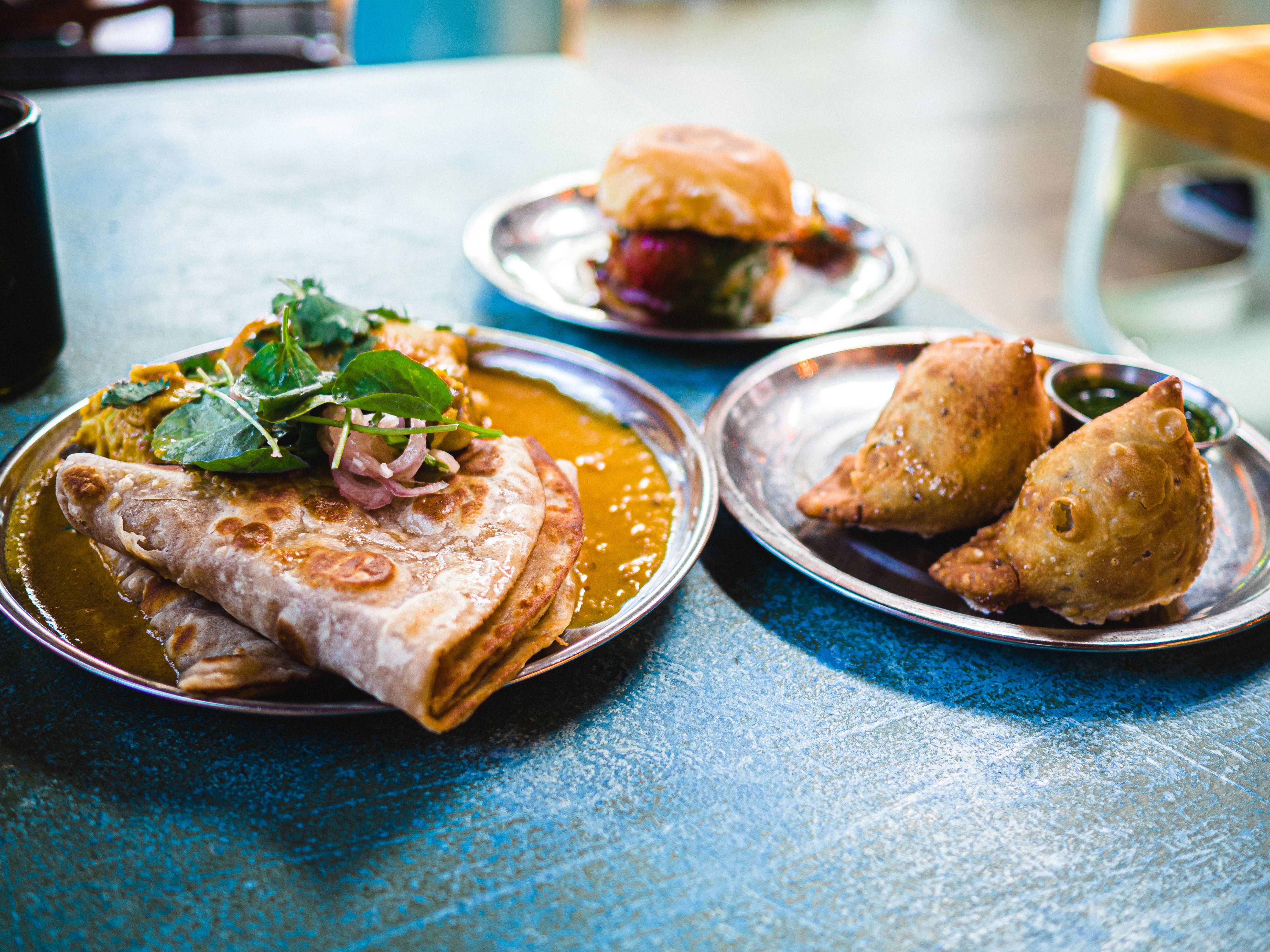 On a blue table there are three plates of food - dal with roti, an empanada, and a hamburger.