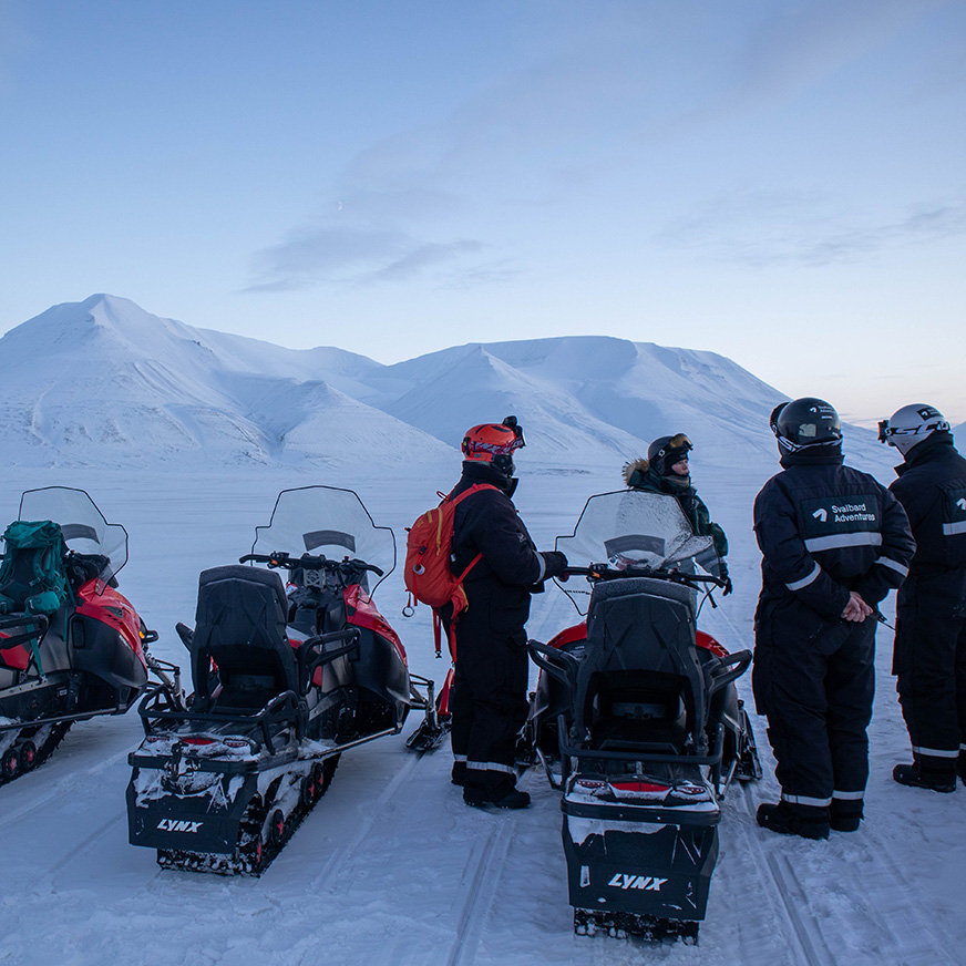 En gruppe står foran snøscootere i et ikledd landskap