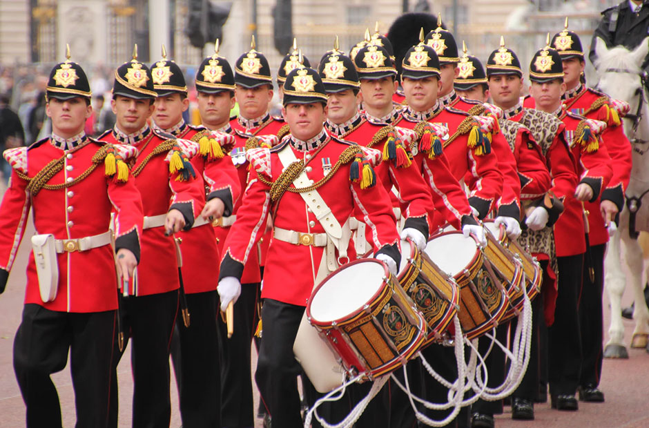 män går på rad iklädda röda uniformsjackor. fyra av dem har trummor. 
